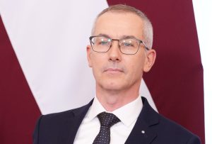 Vīrietis uzvalkā ar brillēm portreta foto uz Latvijas karoga fona.