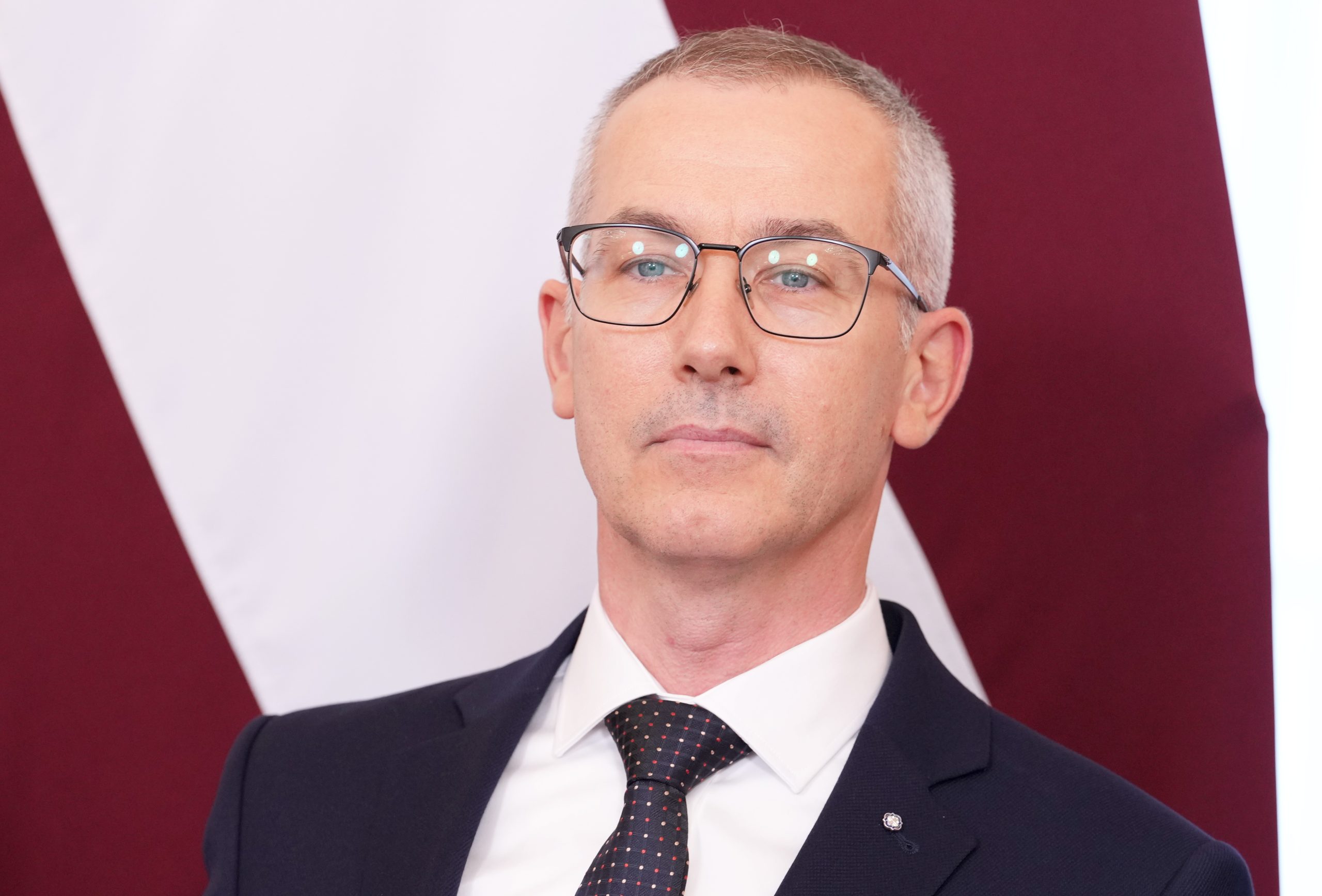 Vīrietis uzvalkā ar brillēm portreta foto uz Latvijas karoga fona.