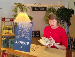 Lasītavā sieviete lasa grāmatu pie galda ar zīmi “Aknīste”