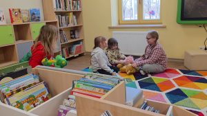 Bērni bērnu bibliotēkas telpā sēž uz paklāja un spēlējas ar rotaļlietām.
