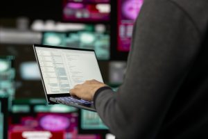 Persona strādā ar portatīvo datoru, ekrānā redzams programmēšanas kods, simbolizējot kiberdrošību un investīciju krāpšanu tiešsaistē.