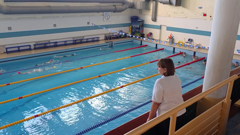 Pārskats uz Jelgavas peldbaseinu, kur trenere vēro peldētāju treniņu