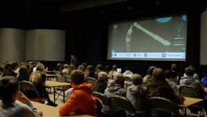Skolēni auditorijā klausās prezentāciju uz lielā ekrāna