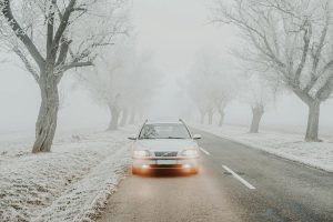 Vecāks Volvo automobilis ar ieslēgtām gaismām brauc pa apledojušu ceļu miglainā ziemas ainavā.