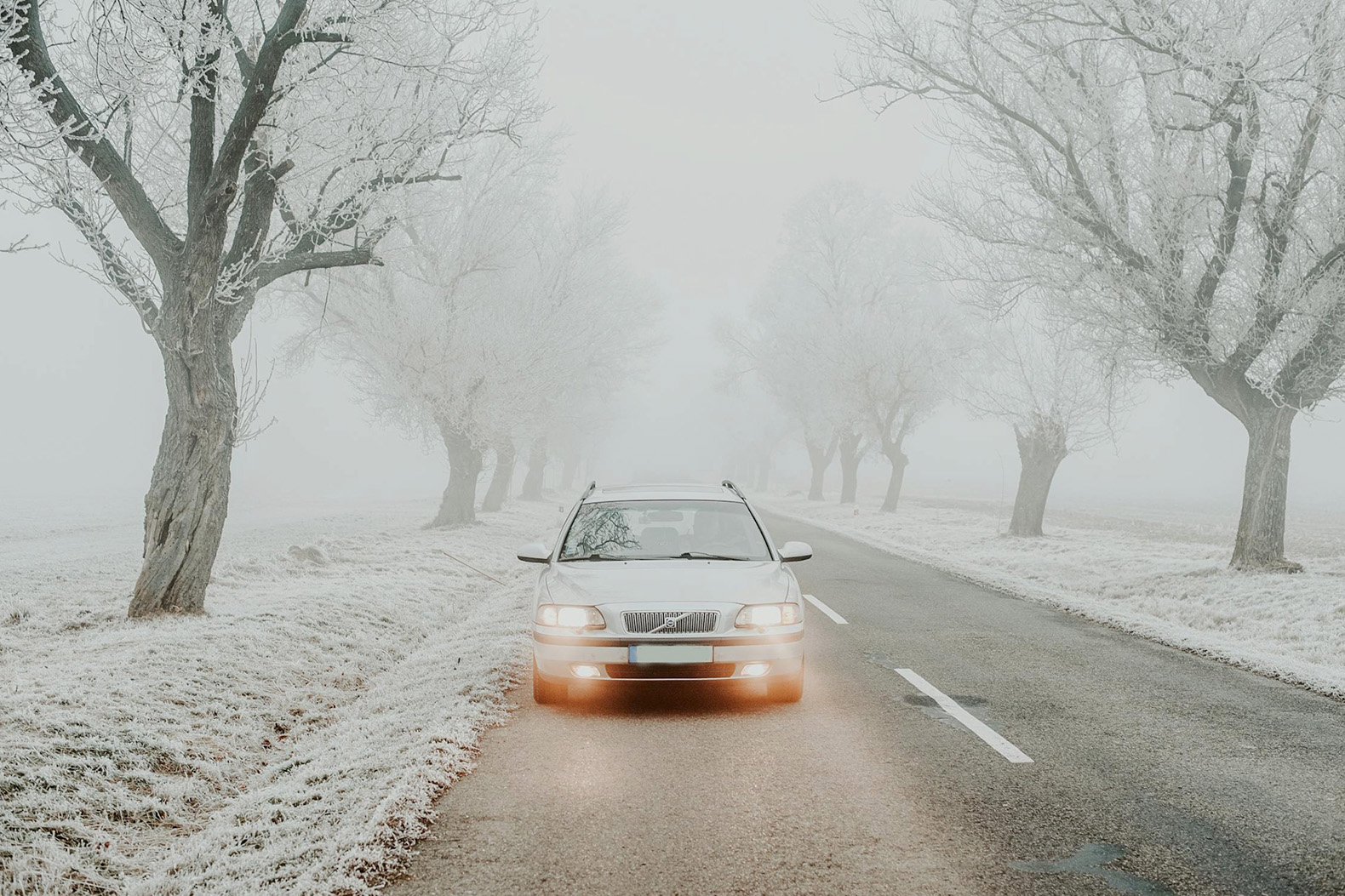 Vecāks Volvo automobilis ar ieslēgtām gaismām brauc pa apledojušu ceļu miglainā ziemas ainavā.