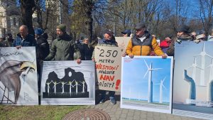 Cilvēki piedalās protesta akcijā pret vēja parku būvniecību, turot plakātus ar ilustrācijām.