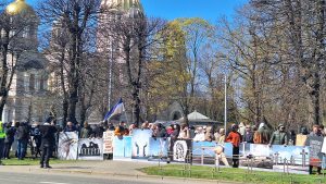Cilvēki piedalās protesta akcijā pret vēja parku projektiem, turot plakātus pie ielas.