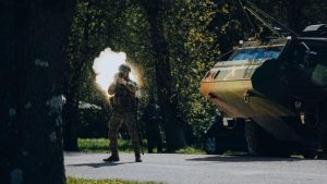 Karavīrs militāro mācību laikā šauj, fonā bruņutehnika meža teritorijā.