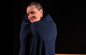 Sieviete uz skatuves ar īsiem matiem un tumšu apmetni