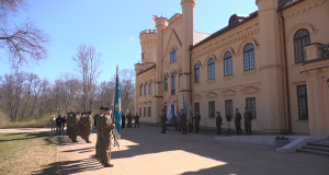 Militārā ceremonija pie vēsturiskas ēkas ar karogiem un karavīru ierindu