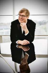 Vīrietis uzvalkā un brillēs sēž pie galda un smaida