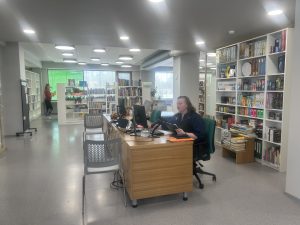 Bibliotēkas darbinieks strādā pie informācijas galda ar datoriem un grāmatplauktiem fonā