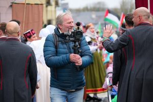Operators filmē pasākumu ar tradicionālo tērpu dalībniekiem un karogiem fonā