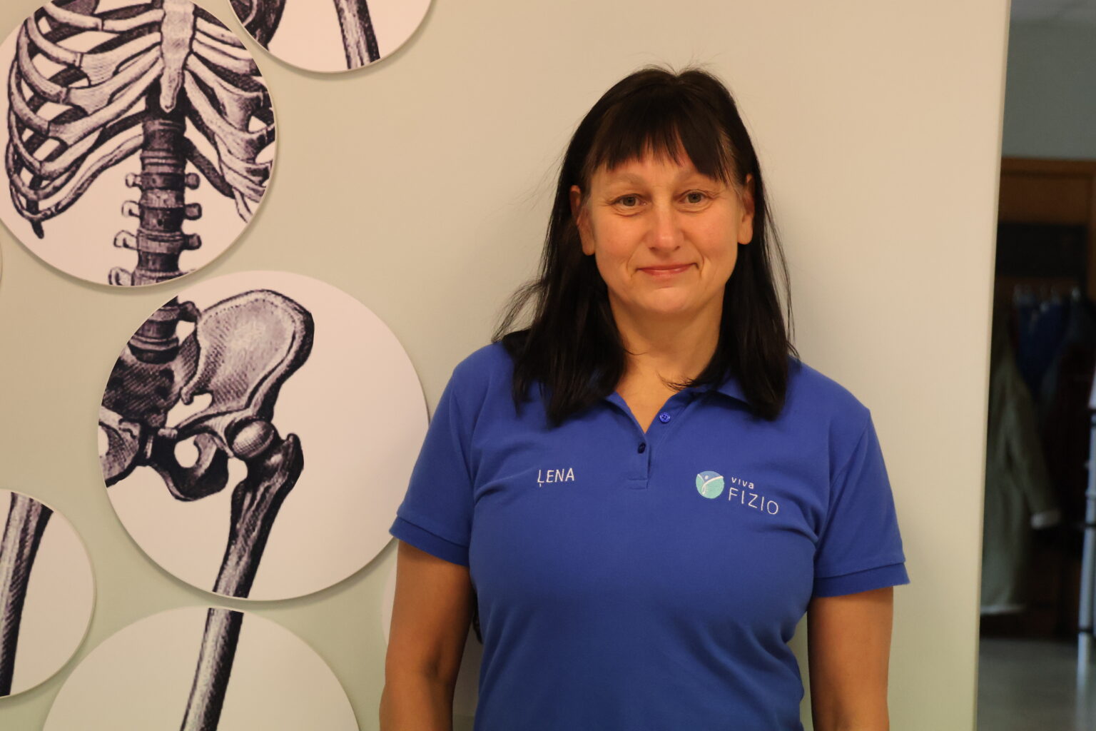 Fizioterapeite Jeļena Miezīte fizioterapijas kabinetā pie sienas ar cilvēka skeleta attēliem