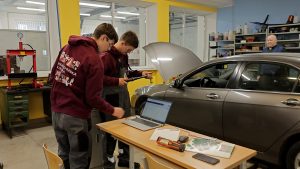 Studenti autoservisa darbnīcā veic diagnostiku pie atvērtas auto motora pārsega