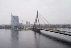 Vanšu tilts Rīgā pār Daugavu miglainā laikā ar pilsētas panorāmu fonā.