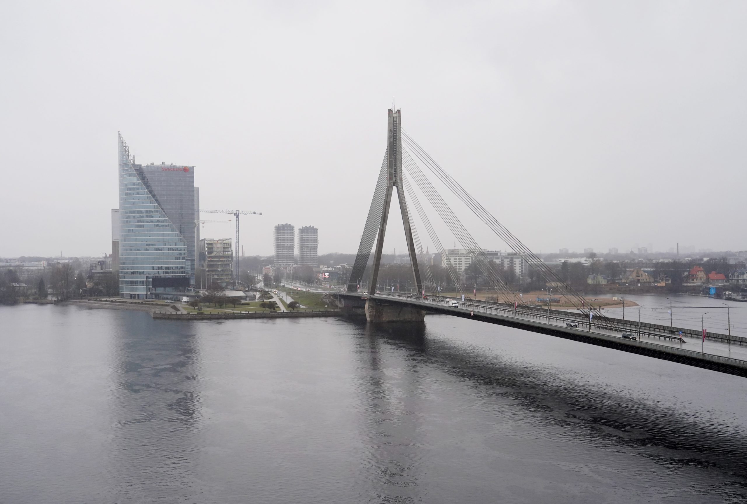 Vanšu tilts Rīgā pār Daugavu miglainā laikā ar pilsētas panorāmu fonā.