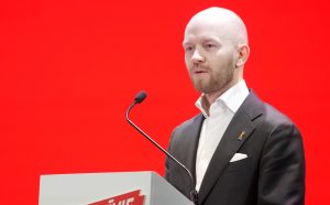 Vīrietis uzstājas pie tribīnes ar mikrofonu uz sarkana fona