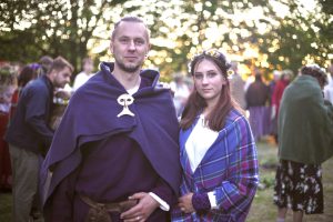 Divi cilvēki svētku tērpos stāv brīvdabas pasākumā pie kokiem