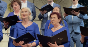 Koris zilā tērpā dzied uz skatuves, turot mapes ar uzrakstu ELPA