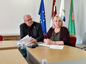 Divi pašvaldības pārstāvji sēž pie galda sanāksmē ar dokumentiem un ieraksta ierīci