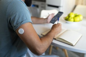 Vīrietis ar glikozes sensoru uz rokas pārbauda datus telefonā un veic pierakstus