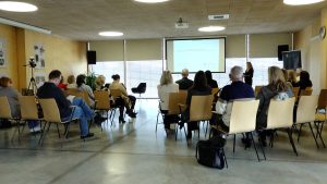 Cilvēki sēž konferenču telpā un klausās prezentāciju uz ekrāna