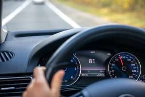 Automašīnas vadītājs brauc pa ceļu, redzams stūres rats un spidometrs