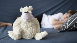 Slimo bērns guļ gultā, priekšplānā plīša rotaļlieta ar sejas masku