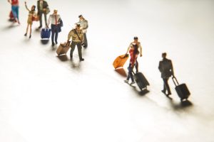 Miniatūru cilvēku figūras ar koferiem simbolizē migrāciju un pārvietošanos.