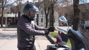 Motociklists melnā ekipējumā stāv pie motocikla un gatavojas braucienam pilsētvidē.