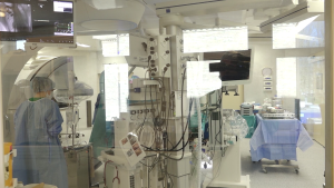 Modernā operāciju zālē medicīnas personāls strādā pie attēldiagnostikas iekārtas un monitoriem.