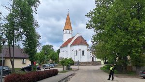 Balta baznīca ciema centrā zem tumšiem lietus mākoņiem