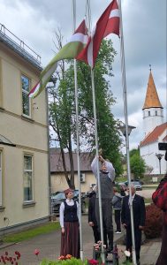 Cilvēki tradicionālos tautastērpos paceļ Latvijas un Sēlijas karogus pie baznīcas