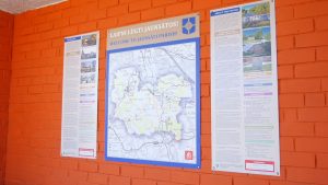 Informācijas stends ar Jaunsātu pagasta karti un aprakstu uz oranžas sienas.