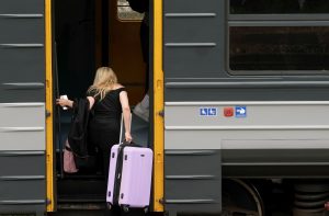 Cilvēks iekāpj vilcienā, turot čemodānu un telefonu pie dzeltenajām durvīm.