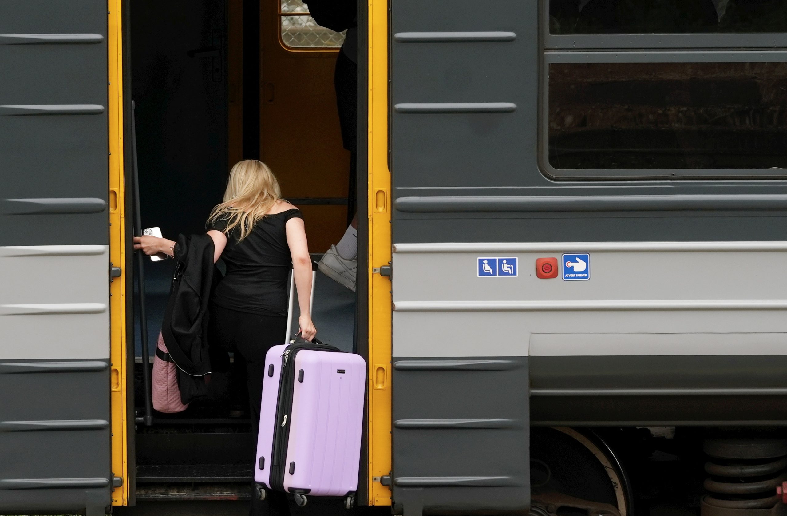 Cilvēks iekāpj vilcienā, turot čemodānu un telefonu pie dzeltenajām durvīm.