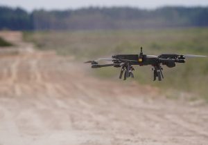 Militārais drons lido virs lauka mācību teritorijā, simbolizējot bezpilota sistēmu nozīmi aizsardzībā.