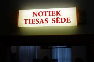 Izgaismota norāde pie tiesas zāles ieejas ar uzrakstu “Notiek tiesas sēde”