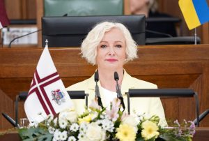 Persona sēž pie tribīnes ar Latvijas un Ukrainas karogiem fonā.