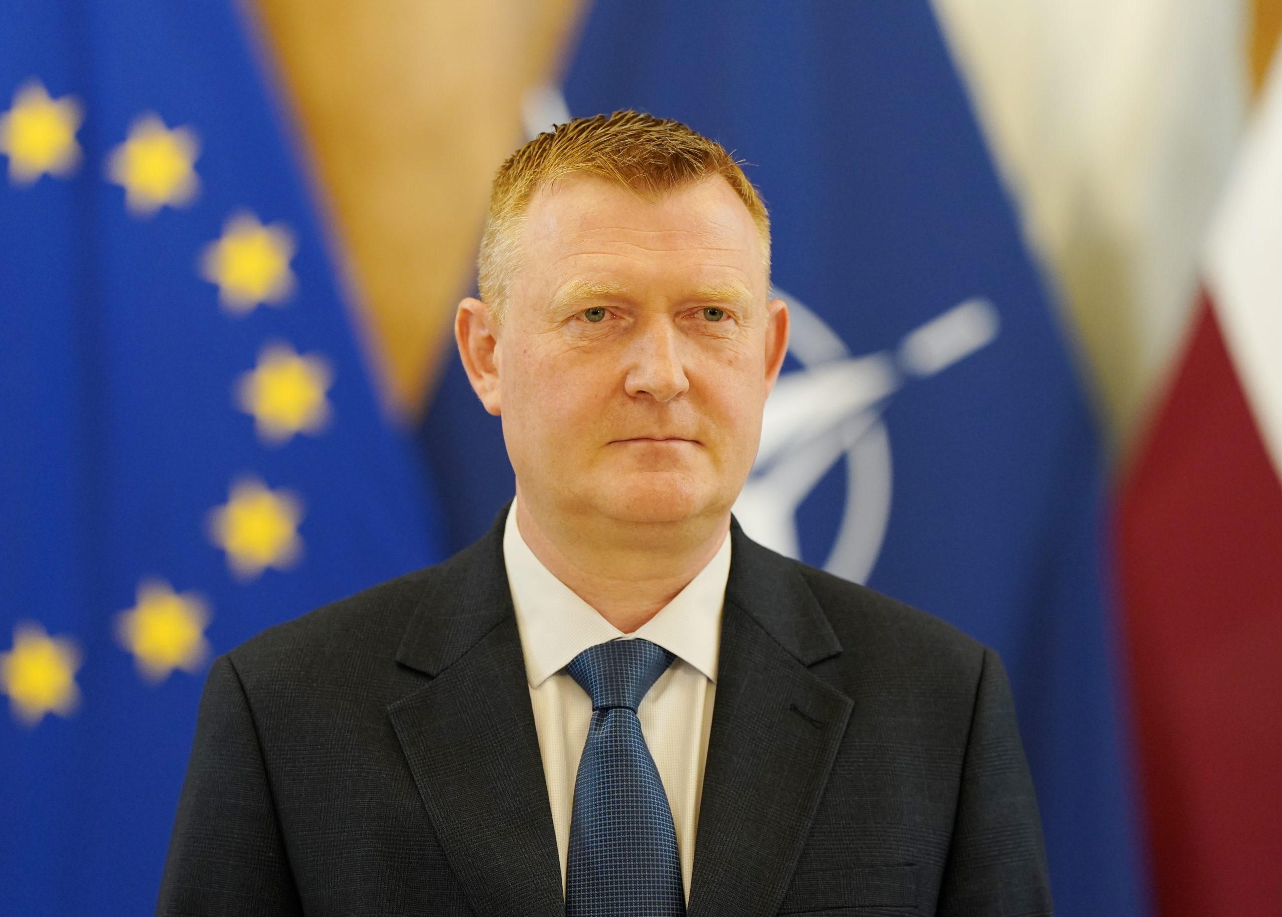 Raivis Melnis stāv uz Latvijas, NATO un Eiropas Savienības karogu fona