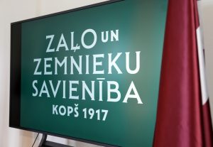Zaļo un Zemnieku savienības logo uz ekrāna blakus Latvijas karogam.