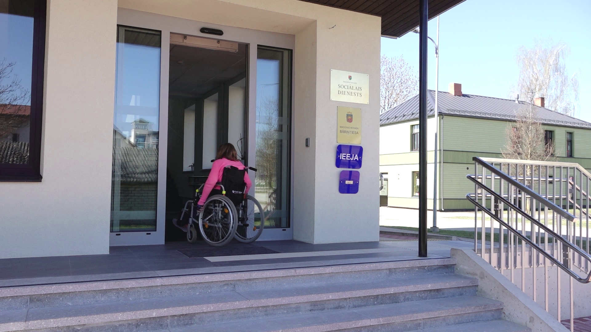 Cilvēks ratiņkrēslā iebrauc sociālā dienesta ēkā pa automātiskajām durvīm.