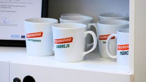 Baltas krūzes ar uzrakstu “Progresīvais” novietotas plauktā