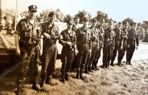 Vīri militārā apģērbā stāv ierindā pie transporta brīvā dabā