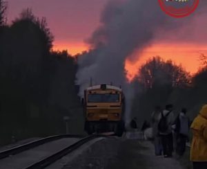 Dīzeļvilciens ar dūmiem uz sliedēm, pasažieri stāv blakus notikuma vietā