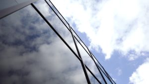 Mākoņu atspulgs stiklotas ēkas fasādē pret zilām debesīm