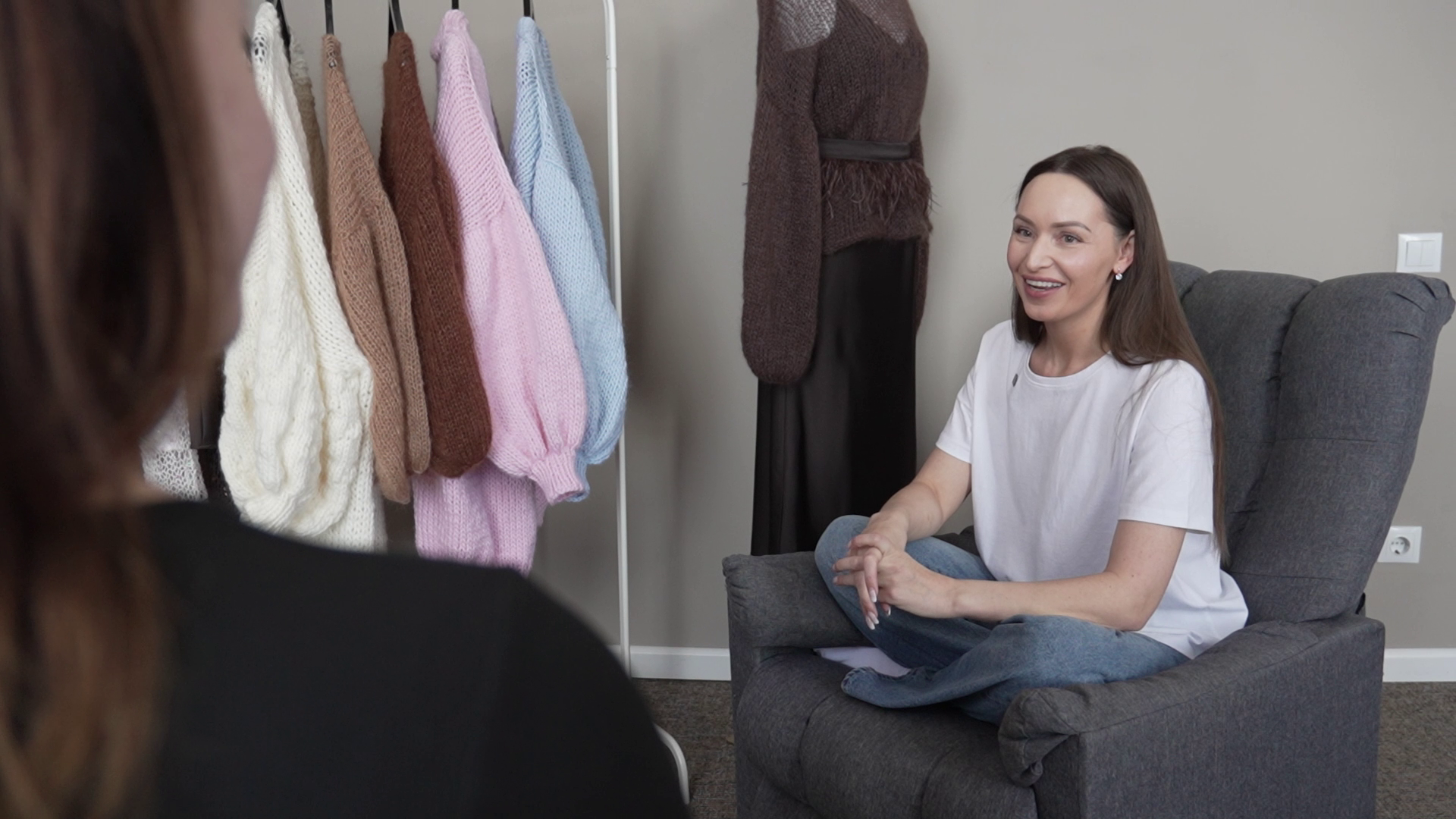 Marita Dinsberga intervijas laikā savā darbnīcā stāsta par adīšanu, fonā redzami viņas darinātie adījumi