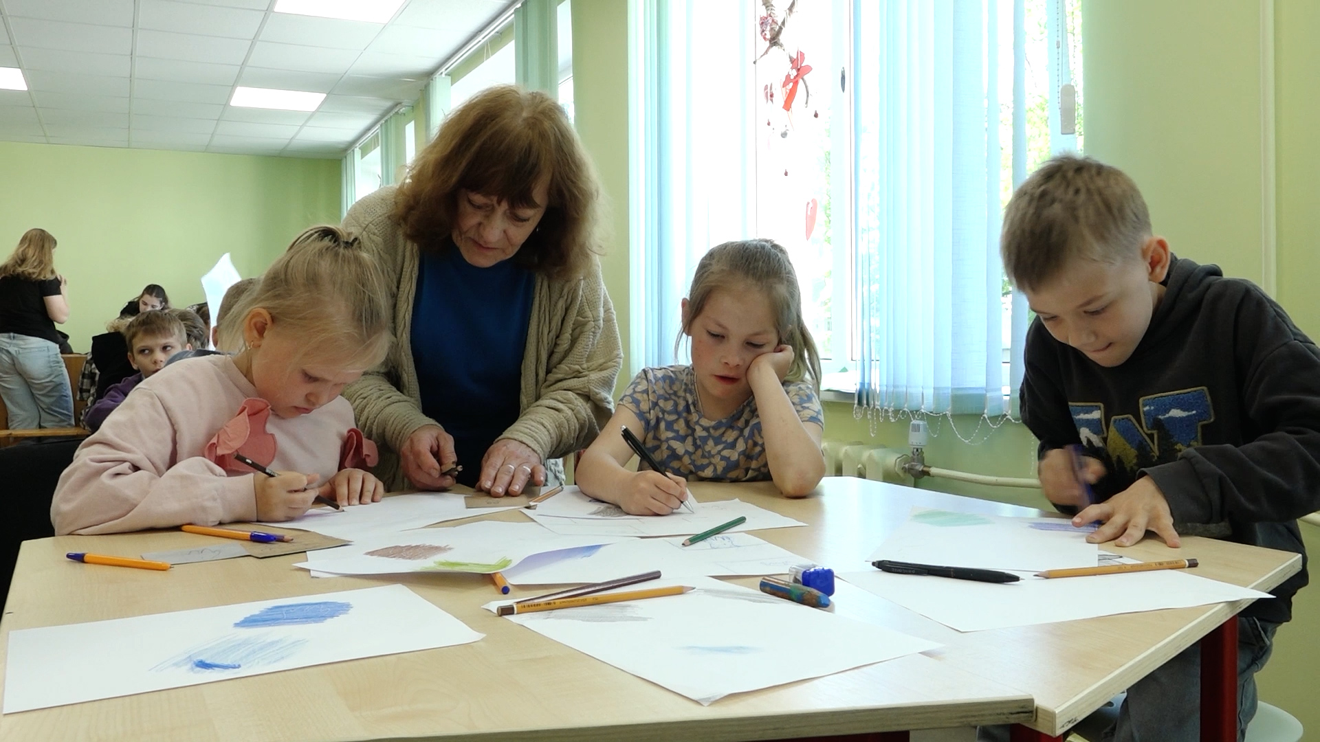 Skolotāja palīdz bērniem radošajā nodarbībā, kamēr skolēni pie galda zīmē un veic uzdevumus uz papīra lapām.