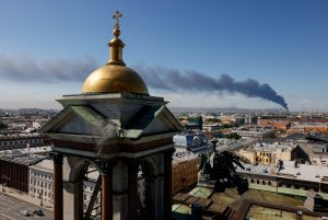 Skats pār pilsētu no ēkas jumta ar baznīcas kupolu priekšplānā un melnu dūmu stabu tālumā virs apbūves.